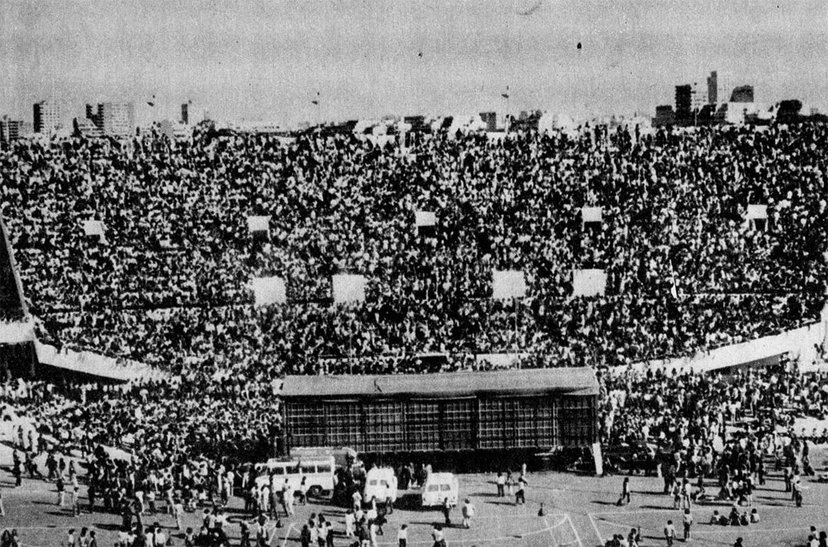 La foto de todos - El fotógrafo se desprendió de la gente, y el sonido se le fue convirtiendo cada vez más en murmullo, llegó a la tribuna vacía que enfrenta a la oficial. Comenzó a subir los primeros 52 escalonés que lo separaban de la cima. Cuando llegó allí sintió un escozor: enfrente y abajo suyo había 15.000 personas absorbidas por la música, como focos el sol implacable destacaba los colores rojos. Las ovaciones las escuchaba como a través de una sordina. Sin ser un profesional, él había captado la foto más monstruosa de B. A. ROCK II. Su asombro fue mayor cuando la reveló: emocionado, a los pocos días la entregó a un redactor de Pelo. El fotógrafo se llama Miguel. Gracias por vernos a todos con tu ojo mecánico. Te envidiamos la idea de llegar hasta allí, pero al menos nos queda tu foto, "la" foto. 62228b25 a60c 4c04 9aeb 5fc4e35027af